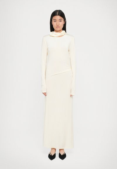 Largo vestido crema con un diseño ajustado, escote alto y detalles fruncidos en un lado, combinado con planos negros de punta.