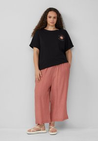 Zwarte katoenen T-shirt met korte mouwen en een zonafbeelding. Losse, roze gekreukte stofbroek. Witte sandalen met bandjes. Neutrale achtergrond.