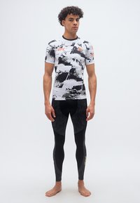 Άνδρας που στέκεται ξυπόλητος φορώντας μαύρο και άσπρο μπλουζάκι συμπίεσης UFC με σχέδιο και μαύρο αθλητικό κολάν με χρυσά λογότυπα.