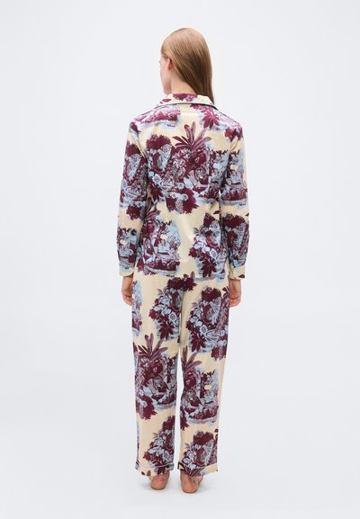 Γυναίκα με ίσια μαλλιά που φοράει ασορτί μακρυμάνικα πουκάμισο και παντελόνι με μωβ και μπλε λουλουδάτο και ζωικό μοτίβο, όρθια ξυπόλυτη με την πλάτη στραμμένη.