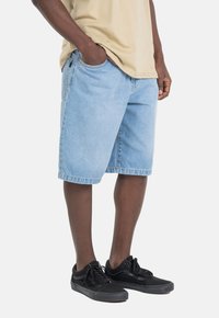 Pantalones cortos de mezclilla azul claro con un ajuste relajado, que cuentan con bolsillos laterales y dobladillos cosidos, combinados con zapatillas de lona negras.