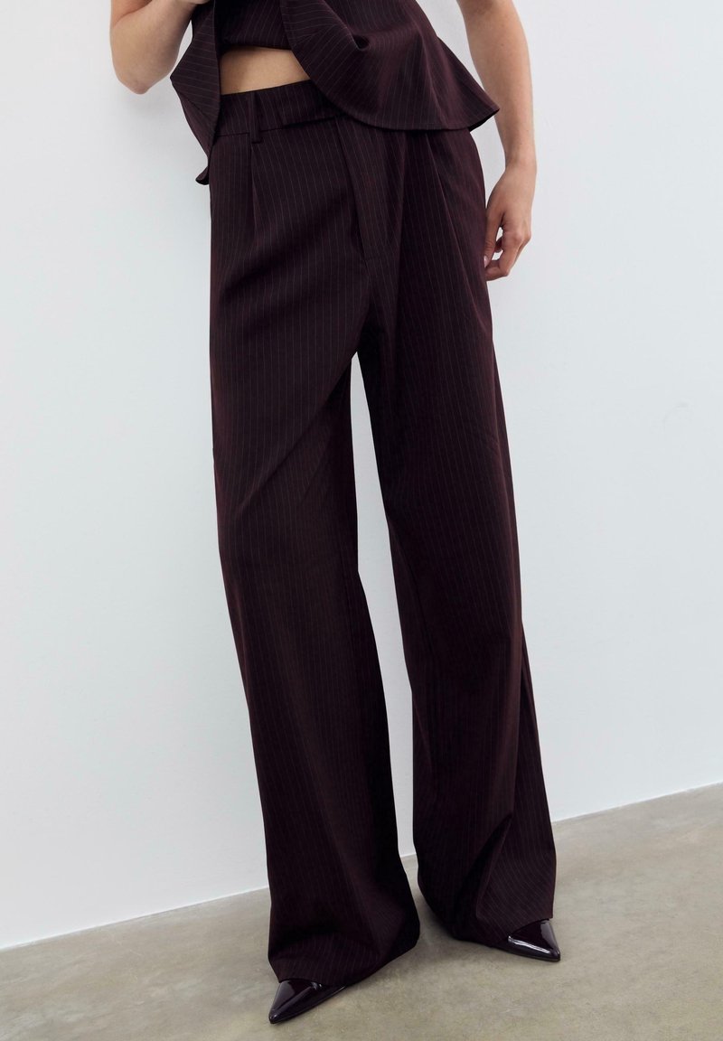 Femme portant un pantalon large à fines rayures foncées avec des chaussures à bouts pointus, debout contre un mur blanc sur un sol en béton.