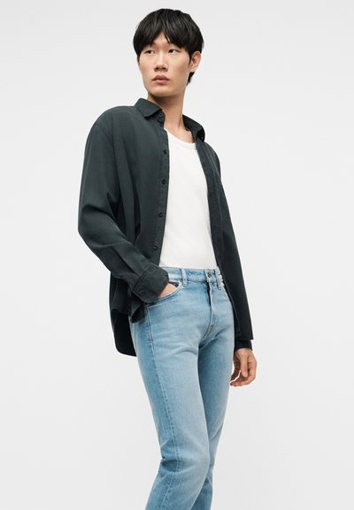 Νεαρός άνδρας που φοράει μαύρο πουκάμισο με κουμπιά πάνω από λευκό t-shirt και ανοιχτό μπλε τζιν, στέκεται με το ένα χέρι στην τσέπη του.