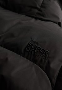 Chaqueta acolchada negra con diseño de acolchado y tejido texturizado, con el logotipo bordado "SUPER DRY JPN" en hilo negro.