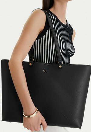 Mujer lleva un bolso grande negro con textura y logo dorado, vistiendo un top sin mangas a rayas negras y blancas y una pulsera dorada en la muñeca.