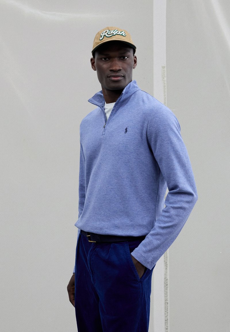 Zils džemperis ar rāvējslēdzēju, izgatavots no mīksta auduma, ar nelielu logo. Kombinēts ar tumšām korduroja biksēm un bēša krāsas cepuri ar zaļu uzrakstu.