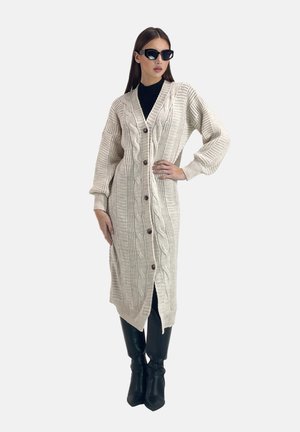 Long gilet crème en tricot texturé, avec un motif torsadé et de grands boutons, associé à un pantalon noir taille haute et des lunettes de soleil.