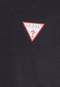 Sort stof med en ribbet tekstur, der har et rødt trekantet logo, der står "GUESS® U.S.A. WASHED JEANS," pyntet med et hvidt spørgsmålstegn.