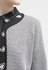 Camicia a righe bianco e nero con una texture geometrica, dotata di un colletto e bottoni, insieme a una linea delle spalle liscia e arrotondata.