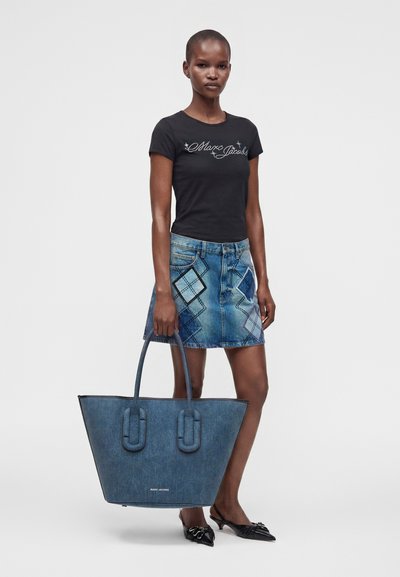 Γυναίκα που φοράει μαύρο t-shirt Marc Jacobs και μπλε τζιν φούστα με patchwork, κρατώντας μεγάλη μπλε τσάντα tote και φοράει μαύρα μυτερά παπούτσια.