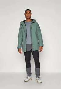 Casaco impermeável verde com capuz, fechos de correr e detalhes pretos. Usado sobre uma camisa cinza com calças escuras que apresentam blocos de cor.