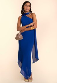 Royalblaues, einseitiges Kleid mit fließendem Design, das einen hohen Schlitz und durchsichtige Textur aufweist. Kombiniert mit goldenen Absatzsandalen und einer beigen Clutch.