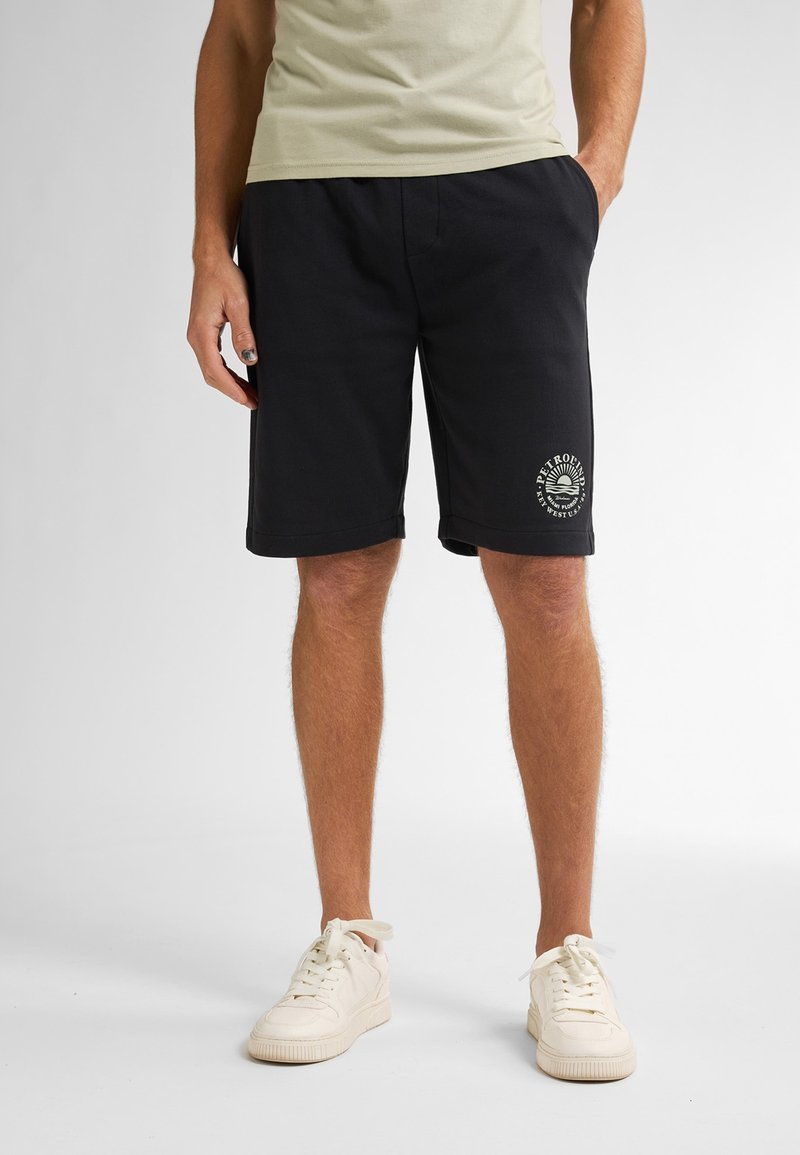 Pantalones cortos de algodón negros con un corte relajado, que presentan un gráfico de logo redondo en la pierna izquierda y bolsillos laterales. Combinados con zapatillas blancas.