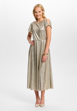 Femme blonde en robe midi dorée brillante à manches courtes, découpe sur une épaule, bracelet et boucles d'oreilles dorés, talons hauts beige, souriante, main sur la hanche.