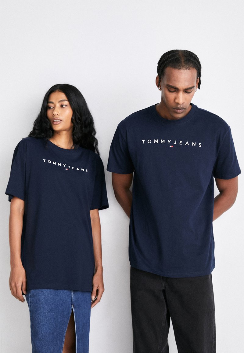 T-shirt di cotone navy con logo "TOMMY JEANS" in bianco, maniche corte e vestibilità rilassata, indossata da due modelli.