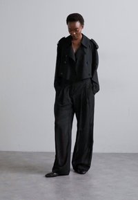 Črna kratka jakna z dvojnimi gumbi, oblečena čez ohlapne črne hlače. Gladka tekstura in minimalističen dizajn, brez vzorcev ali dodatkov.