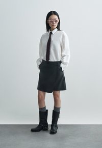 Bílá košile na knoflíky, tmavá kravata, černá kožená zavinovací sukně, šedé podkolenky a černé kotníkové boty s řetízkovým detailem. Světlé pozadí.