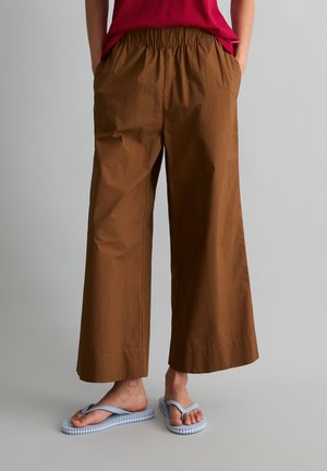 Osoba ubrana w luźne brązowe spodnie do połowy łydki, czerwony top i jasnoniebieskie klapki, stojąca na jednolitym tle.