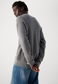Junger Mann mit dunkler Haut und kurzen Dreadlocks, der einen grauen Strickpullover und blaue Jeans trägt und mit hinter dem Rücken verschränkten Händen steht.