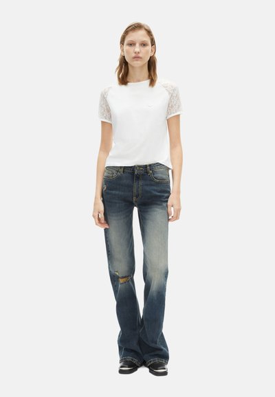 Witte t-shirt met kanten mouwen, gecombineerd met licht versleten blauwe jeans. De jeans hebben een rechte pijp en subtiele vervaging.