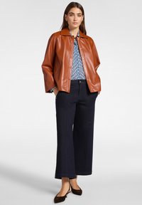 Donna con giacca di pelle marrone, blusa blu con motivo, pantaloni a gamba larga scuri e décolleté neri con slingback, in piedi con le mani nelle tasche.