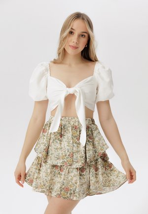 Top alb cu croială scurtă, manșete bufante și detaliu cu nod în față, asortat cu o fustă florală cu volane în culori delicate. Țesătură texturată.