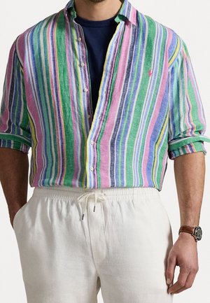 Mann trägt ein mehrfarbiges, vertikal gestreiftes Hemd über einem marineblauen T-Shirt und weißen Hosen mit Kordelzug, dazu eine braune Armbanduhr.