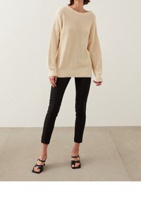 Suéter de punto color crema con cuello ancho y mangas largas, combinado con pantalones ajustados negros y sandalias de tacón negras.