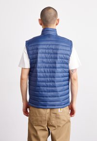Vesta puffer fără mâneci de culoare albastră, cu textură matlasată și guler înalt, purtată peste un tricou alb. Include un detaliu cu logo pe spate.