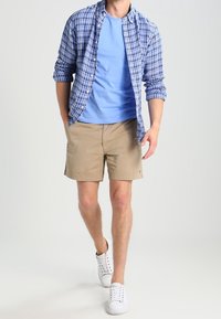 Modrá kostkovaná košile přes světle modré tričko, khaki šortky a bílé tenisky. Šortky jsou do kolen; košile má vyhrnuté rukávy.