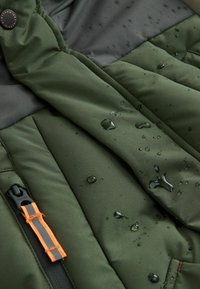 Nepremokavá zelená puffa bunda s čiernymi akcentmi, na látke sú kvapky vody a je vybavená oranžovou zipsovou ťahadlom s reflexným prúžkom.