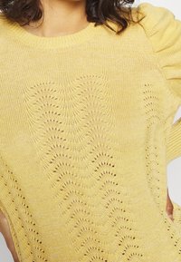 Pull-over en maille jaune pastel avec un motif ondulé à mailles ajourées, un col rond et des manches légèrement bouffantes. Finition texturée.