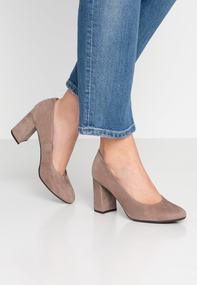 Personne portant des escarpins en daim taupe à talons blocs associés à un jean en denim bleu raccourci sur un fond uni.