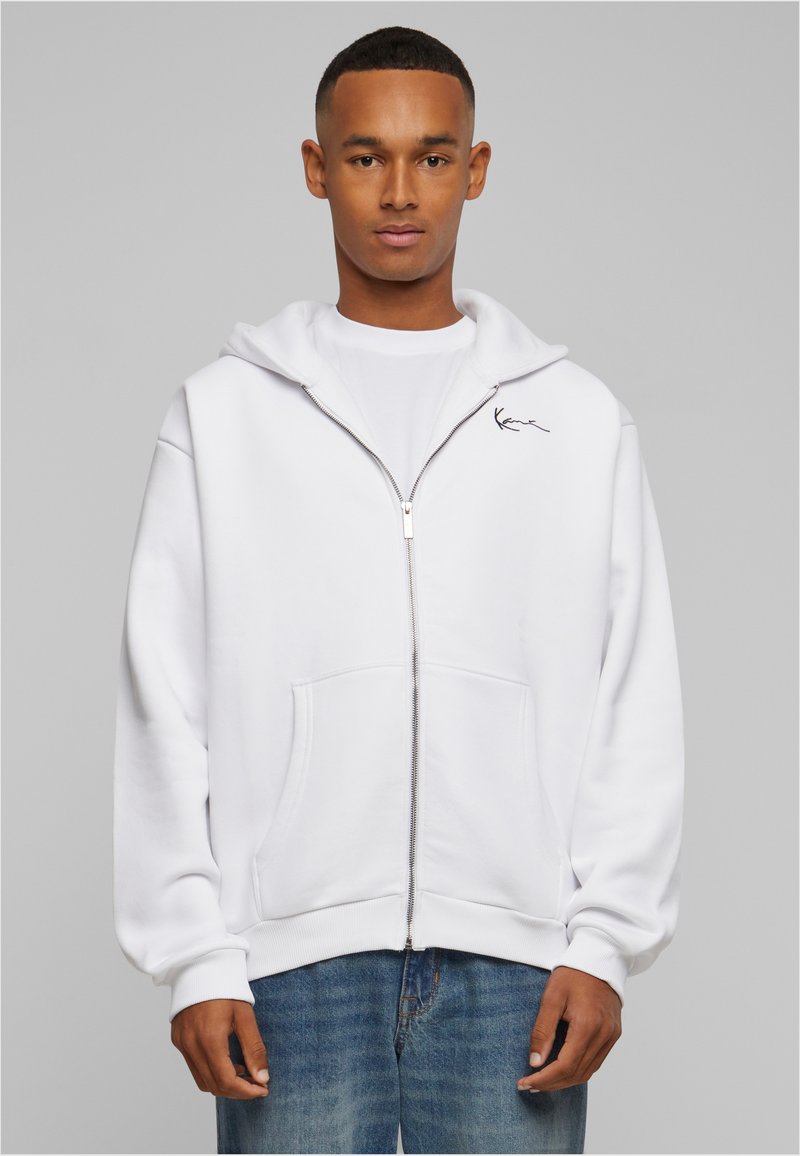 Jonge man draagt een witte zip-up hoodie met voorzakken en een klein zwart logo op de linkerborst, gecombineerd met een blauwe spijkerbroek.
