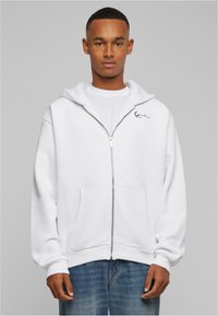 White zip-up hoodie made of soft fabric, featuring a kangaroo pocket and a small embroidered logo on the chest. Long sleeves with ribbed cuffs.