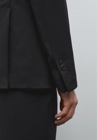 Czarna marynarka o tailoringu z gładką fakturą, z dwoma guzikami na mankiecie i wyraźnie zarysowanym krojem. Materiał jest lekki i elegancki.