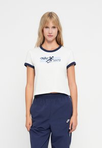 Balta kokvilnas piegriezuma topiņš ar tumši zilu ribota kakla un piedurkņu apdari. Uz topiņa ir "Nike 1972" logo zilo grafisko druku. Apvienots ar tumši ziliem sporta biksēm.