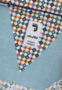 Multikleurige stoffen met bloemmotieven op een witte achtergrond, gecombineerd met een gestructureerde blauwe stof. Etiket met de tekst "J&JOY, Ontworpen in België."