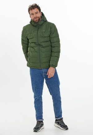 Grön puffjacka med huva, hög krage och kviltad yta, bärs med blå jeans och svarta sneakers, står framför en vit bakgrund.