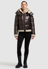 Braune gepolsterte Jacke mit einer Kunstschafswollkapuze und -verzierung, ausgestattet mit einem Reißverschluss und Klappentaschen sowie schwarzen Akzenten. Kombiniert mit schwarzen Leggings und Stiefeln.