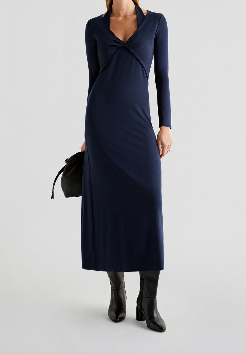 Femme portant une longue robe bleu marine avec un col torsadé, des bottes à talons noires et tenant un petit sac à main noir.