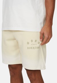 Pantaloni corti color crema in un tessuto morbido, con un design testuale sulla coscia sinistra in grigio chiaro. Vestibilità dritta con una superficie liscia.