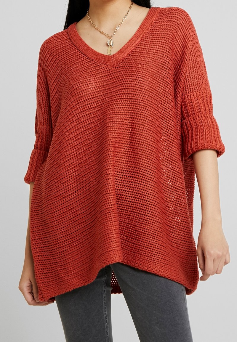 Femme portant un pull ample en maille orange brûlé à col en V avec les manches retroussées et un jean gris foncé, posant devant un fond gris uni.