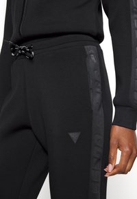 Svart joggebukse med strammesnor i midjen, sidelomme og teksturert logo-panel på leggen. Glatt stoff med en tettsittende design.