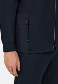 Giacca sportiva blu navy con chiusura frontale a zip, dotata di tasche laterali, texture liscia e taglio aderente. Dettaglio distintivo vicino all'orlo.