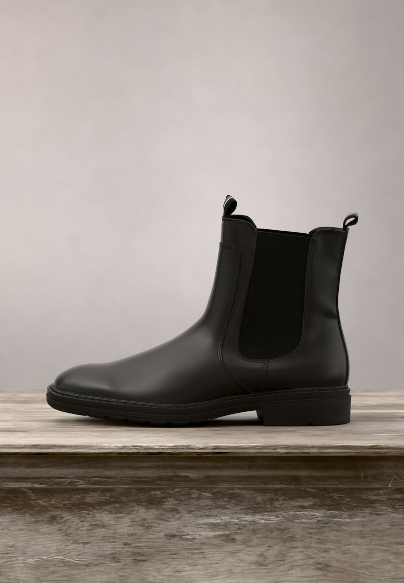 Bottine Chelsea en cuir noir avec panneaux latéraux élastiques et languettes, exposée sur une surface en bois sur un fond neutre.