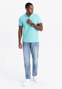 Tyrkysová polo košile s límcem, kombinovaná se světle modrými džíny a bílými teniskami. Výrazné náramkové hodinky na levém zápěstí. Jednoduchý design.