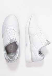 White athletic shoes with a mesh upper, hexagonal pattern, grey accents, padded collar, and flat laces. Sole is textured for grip.