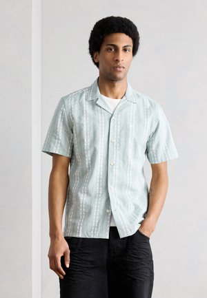 Άνδρας με afro χτένισμα φορά ανοιχτό μπλε κοντομάνικο πουκάμισο με κάθετες λευκές ρίγες και μαύρο παντελόνι, στέκεται μπροστά σε απλό φόντο.