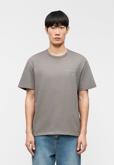Jeune homme aux cheveux noirs raides portant un t-shirt gris avec le logo Hogan et un jean bleu, se tenant devant un fond blanc uni.
